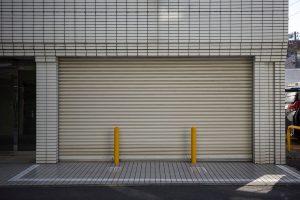 Closed beige roller shutter door between white tiled pillars with two yellow bollards in front