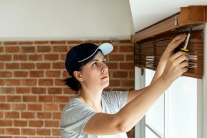 Woman in navy cap uses screwdriver on wooden blind headrail by red brick wall