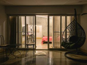 Dim room with open glass sliding doors revealing a lit bedroom, black hanging egg chair and bistro table