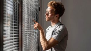Curly-haired man wearing round glasses parts white blind slats with a finger in sunlight