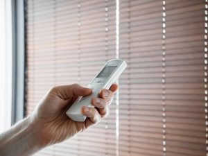 Hand holding a white remote control with a small display toward closed horizontal brown slat blinds
