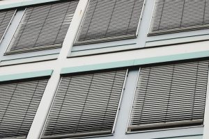 Building facade with large windows covered by gray horizontal slat blinds, framed by white walls and pale green trim