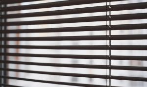 Close-up of dark brown horizontal slat blinds with thin ladder cords, backlit by blurred buildings outside