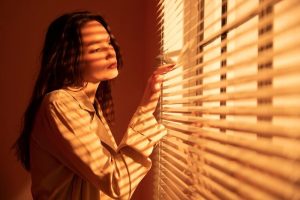 Woman with long dark hair parts horizontal slatted blinds with her fingers, warm orange light striping her face