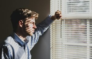 Man in blue shirt leans on wall as sunlight through window blinds stripes his face