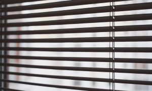 Close-up of dark brown horizontal slat blinds with vertical lift cords, light filtering through blurred buildings outside